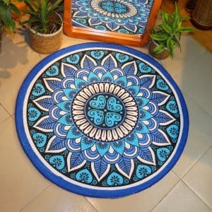 Handwoven round jute and cotton floor mat with colorful mandala design placed on tiled floor, surrounded by woven baskets and green plants